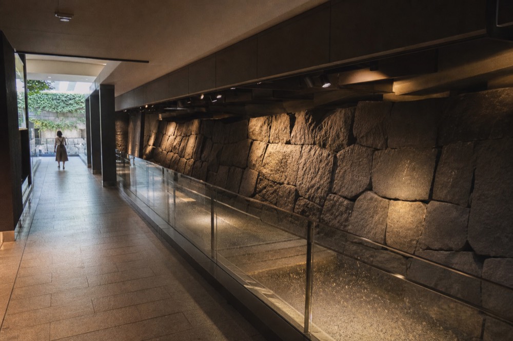 A dark underground exhibition hall with a long stone wall lit by warm spotlights