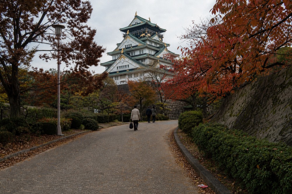 A tall castle keep with layered white walls and green roof tiles seen from a park pathway