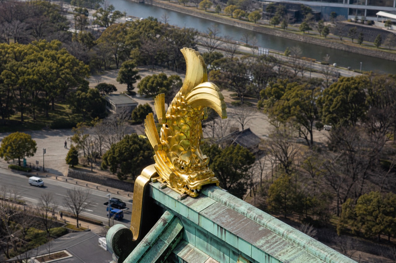 A close-up of the golden shachihoko on the roof of Osaka Castle, with the green copper roof tiles and blue sky behind it