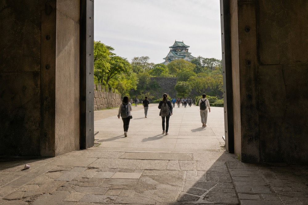 Visitors with small daypacks walking toward a castle gate across a stone plaza