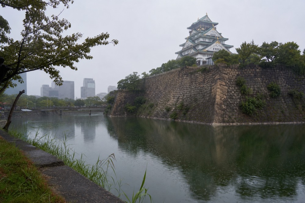 Grey rainy-season clouds over the Osaka Castle tower seen from the outer moat, with reflections on wet water