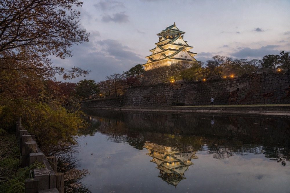 A castle keep reflected in still moat water at dusk with floodlights beginning to glow