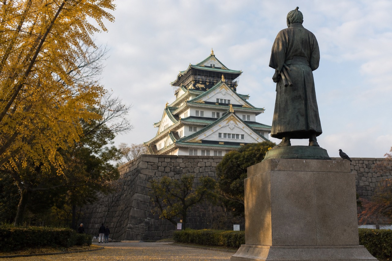A statue of a samurai commander stands before the white walls and green roofs of Osaka Castle's tower