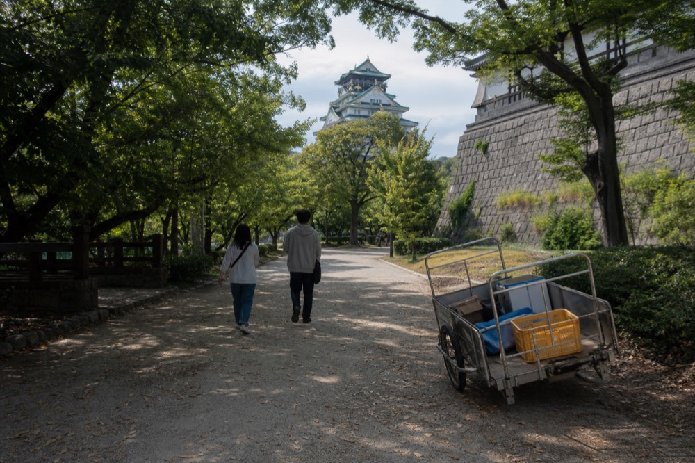 A tree-lined path through Osaka Castle Park leading toward the tower