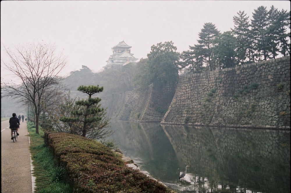 A wide shot of the outer moat and the stone walls reflecting on the water with the castle tower visible behind the trees