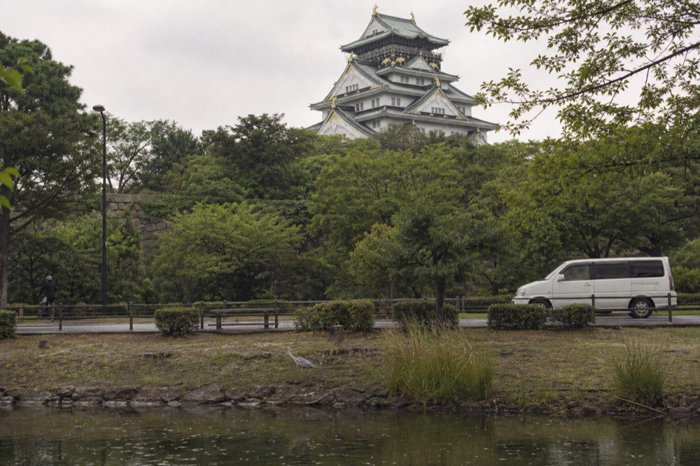 The white Main Tower of Osaka Castle seen from the outer moat with green trees in the foreground