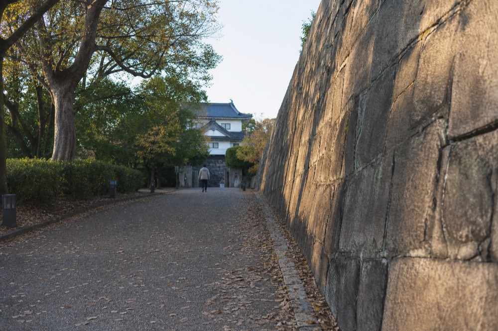 A stone gate and wall along a tree-lined path in a castle park in late afternoon light