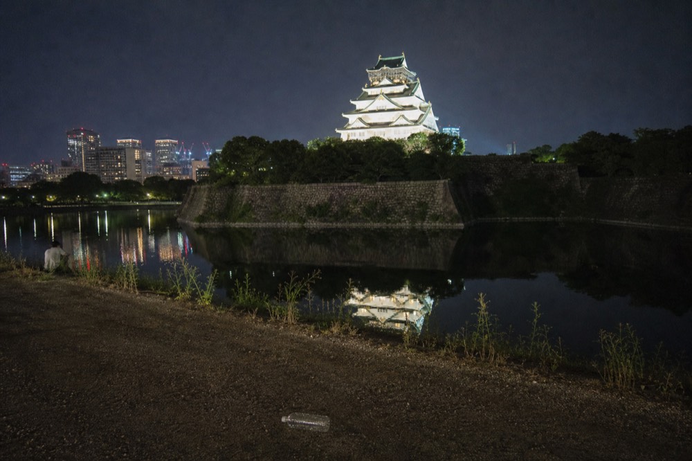 A wide view of the outer moat at night, with the illuminated tower reflected in the still water and city lights in the distance